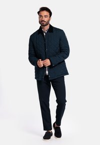 Gepatchworkte donkerblauwe jas met een zwarte kraag, voorzien van drukknoopjes en twee zijzakken, gedragen over een plaid overhemd en donkere broek.