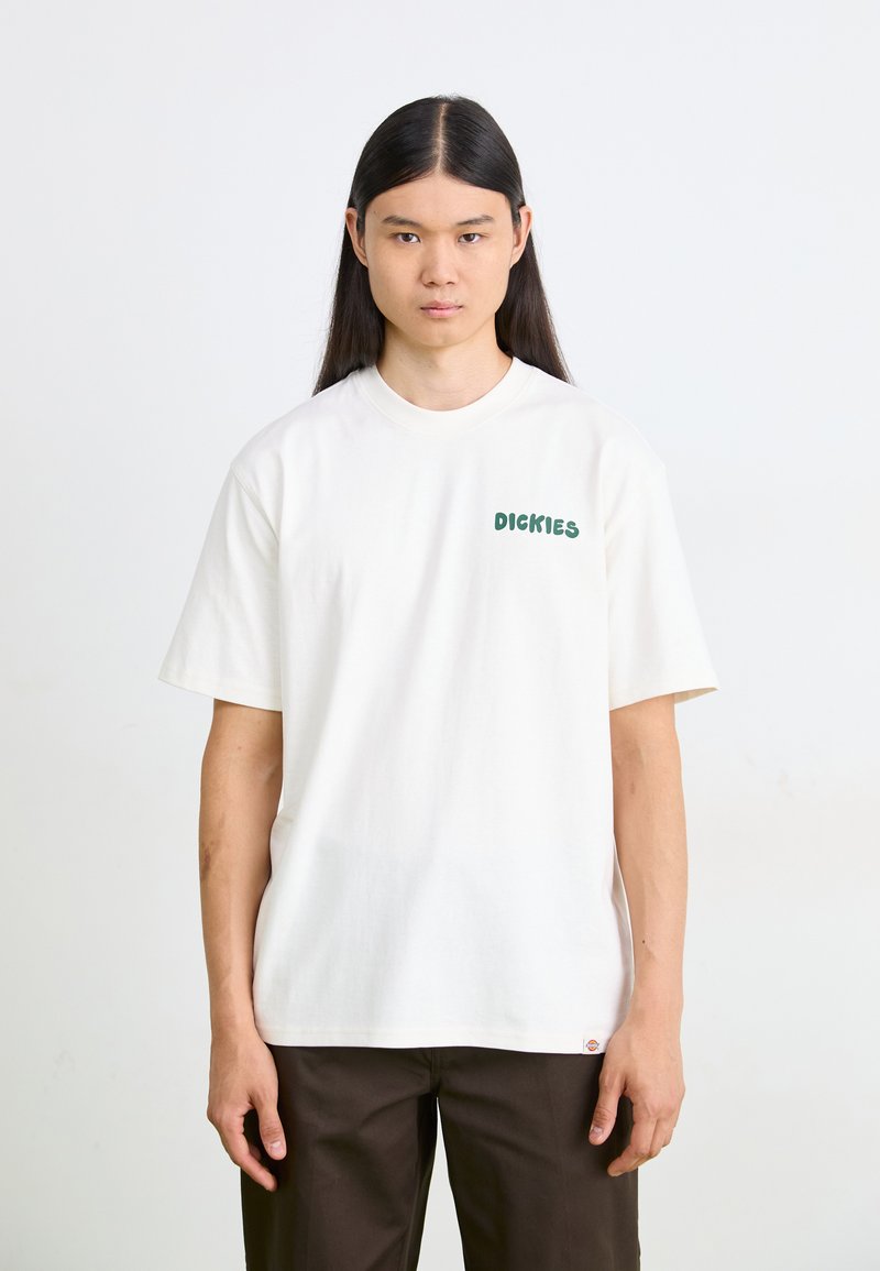 T-shirt bianco in cotone con maniche corte, colletto tondo e testo verde "DICKIES" sul lato sinistro del petto. Abbinato a pantaloni marrone scuro.