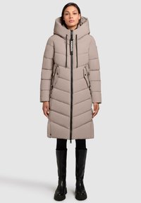 Lange beige pufferjas met een chevronpatroon, grote capuchon, zwarte rits en twee zijzakken met rits. Gecombineerd met zwarte knie hoge laarzen.