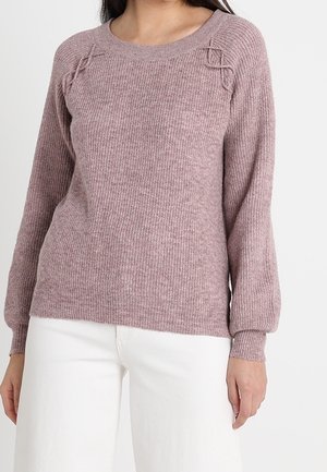 Femme portant un pull en maille mauve avec des détails torsadés près des épaules et un pantalon blanc, debout devant un fond blanc uni.