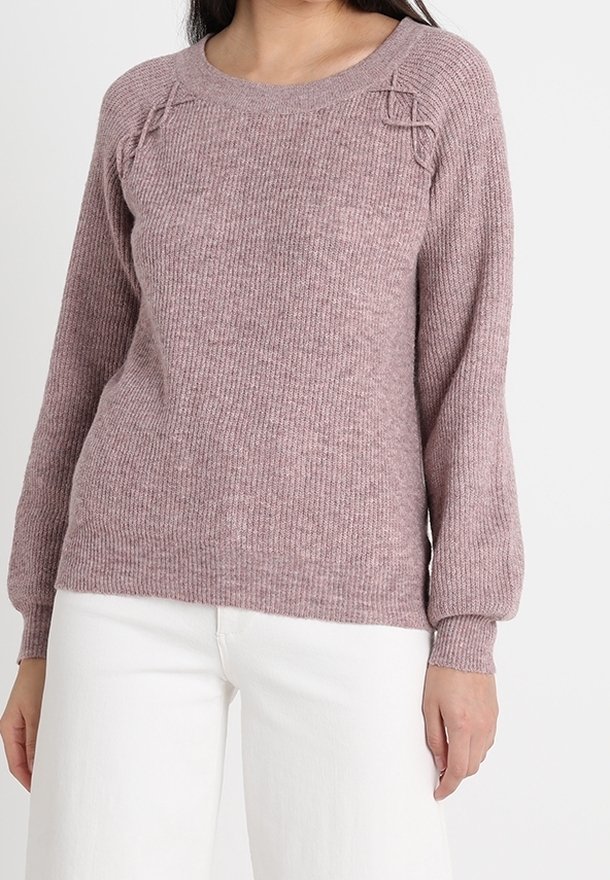 Femme portant un pull en maille mauve avec des détails torsadés près des épaules et un pantalon blanc, debout devant un fond blanc uni.