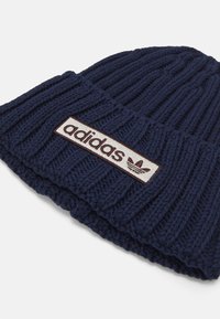 Σκούφος από μαλλί σε ναυτικό μπλε με ριγωτή υφή, διαθέτει ορθογώνια λευκή επιφάνεια με το λογότυπο και το έμβλημα της "adidas" σε μπορντώ.