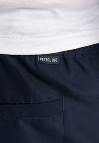 Tissu bleu marine à texture lisse avec une poche visible et une étiquette noire portant l'inscription "PETROL IND" cousue près de la ceinture.