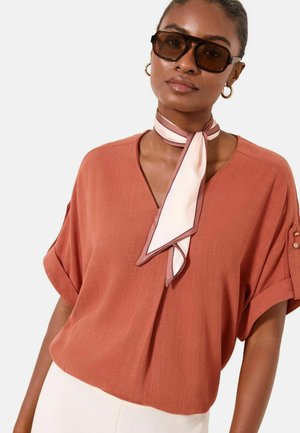 Frau mit rostfarbener V-Ausschnitt-Bluse, hellrosa Schal, der am Hals gebunden ist, großen braunen Sonnenbrillen und goldenen Creolen vor weißem Hintergrund.