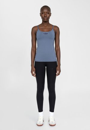 Femme debout, face à l'avant, portant un haut bleu sans manches, des leggings noirs, des chaussettes blanches et des baskets blanches sur un fond uni.