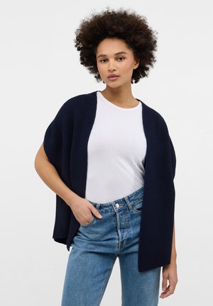 Navy gestrickte Strickjacke mit offenem Vorderteil, kurzen Ärmeln und geripptem Muster, getragen über einem weißen T-Shirt und kombiniert mit hellblauen Jeans.