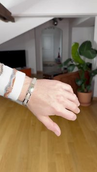 Narukvica od srebrnog i zlatnog metala s teksturiranim dizajnom, koja sadrži međusobno povezane karike. Nosena na ruci nasuprot pozadini drvenog poda.