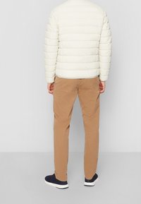 Giubbotto imbottito beige chiaro con cuciture orizzontali, abbinato a chinos color sabbia e sneaker navy. Texture liscia, silhouette casual.