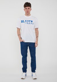 T-shirt bianca a maniche corte con testo blu "BLAUER", abbinata a jeans in denim blu e sneakers bianche con accenti blu.