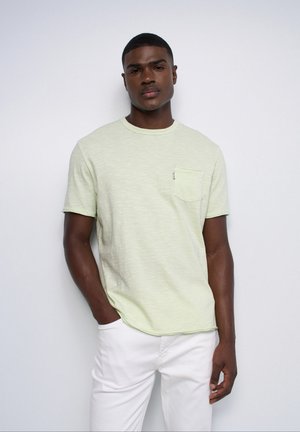 Young man with short hair wearing a light green short-sleeve t-shirt and white pants, standing with one hand in pocket against a plain background.