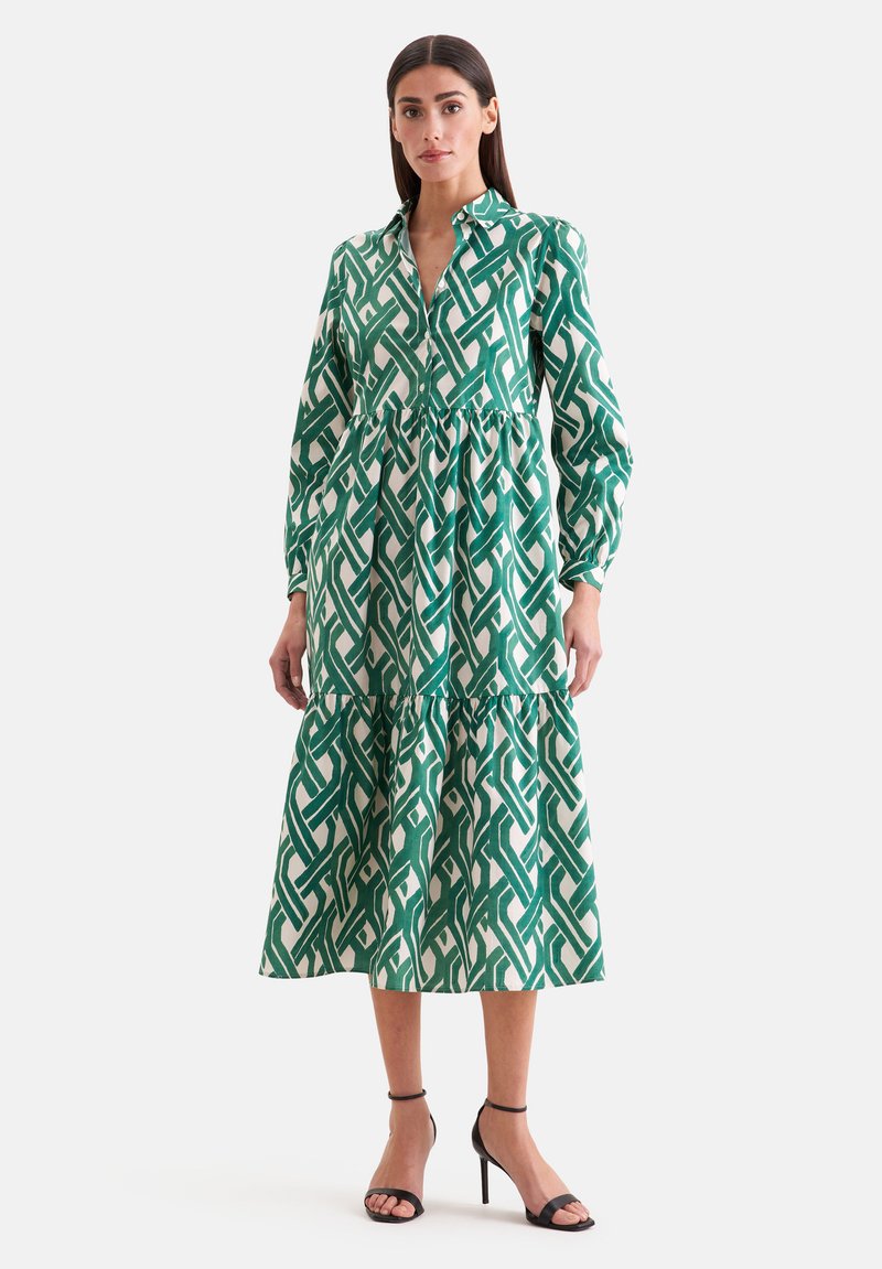 Femme portant une robe midi à manches longues, boutonnée, à motif vert et blanc avec des volants en étages, associée à des talons ouverts noirs.