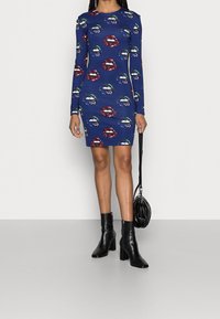 Long-sleeve blue dress with repeated lip graphic prints. Fitted design, mid-thigh length, paired with black heeled ankle boots and a small bag.