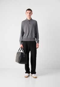 Polo grigio a maniche lunghe con tre bottoni, pantaloni neri, tiene in mano una borsa in pelle nera con bordi arrotondati e doppi manici. Sneakers bianche.