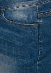 Džínové kalhoty ve středně modré barvě, s zadní kapsou a černým nýtem. Textura je mírně vybledlá, vykazuje známky nošení.