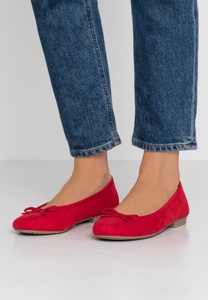 Person trägt rote Ballerinas mit kleinen Schleifen und blaue verkürzte Jeans und steht auf einer hellen Oberfläche.