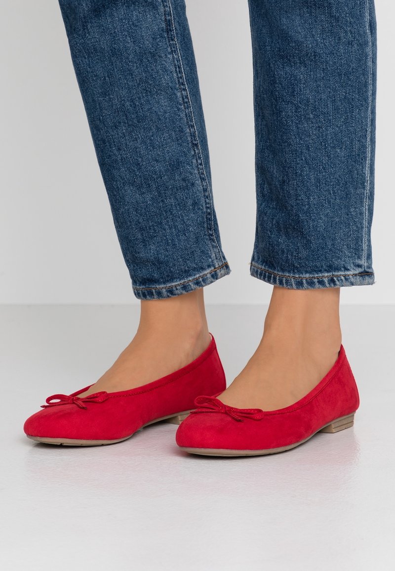 Personne portant des ballerines rouges avec de petits nœuds et un jean bleu coupé debout sur une surface claire.