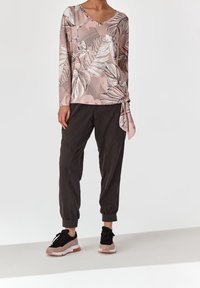 Langärmlige pinke Bluse mit weißem und grauem Blättermuster, mit V-Ausschnitt und seitlichem Bindeband, kombiniert mit dunklen, schmal geschnittenen Hosen und schwarzen Sneakers.