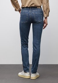 Jeans ajustados de mezclilla azul oscura con una textura suave, que presentan dos bolsillos traseros y detalles en el panel lateral. Combinados con zapatillas blancas.