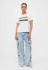 T-shirt en coton blanc avec une rayure horizontale en imprimé léopard, associé à un pantalon cargo bleu clair avec plusieurs poches et des jambes larges.