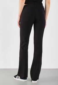 Black dress pants with a straight leg design, waistband with loops, and subtle rear pockets. Worn with dark sneakers and a black top.