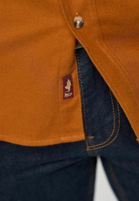 Oranje denim shirt met een knoopsluiting, subtiele textuur en een klein paars en goud logo-label op de naad, gecombineerd met donkere jeans.