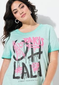 Mintegrøn kortærmet T-shirt med sort og pink graffiti-inspireret tekst, herunder "PARIS" samt designelementer som en krone og diamant.