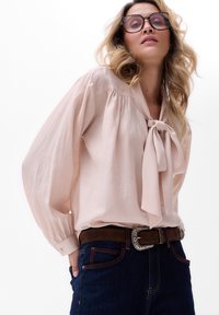 Jasnoróżowa bluzka z wiązaniem przy dekolcie, obszerne rękawy i teksturowana tkanina. Noszona z ciemnoniebieskimi dżinsami i brązowym paskiem.