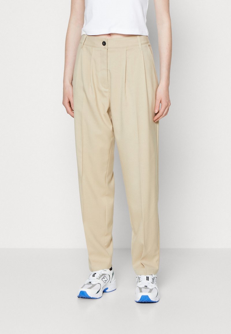 Beige geplooide broeken met een vlakke taille, voorzien van een knoopsluiting bij de tailleband. Lichtgewicht, gladde stof, gecombineerd met wit en blauwe sneakers.