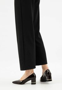 Scarpe con tacco alto in pelle verniciata nera con punta affusolata e tacco largo, dotate di una fascia decorativa dorata. Indossate con pantaloni neri larghi.