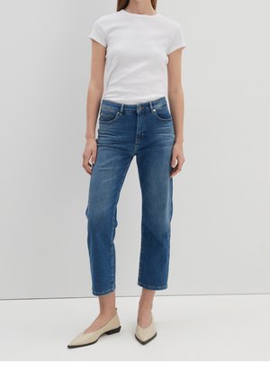 Kvinde iført en hvid T-shirt med korte ærmer, blå højtaljede korte jeans og beige spidse flade sko, stående mod en lys baggrund.
