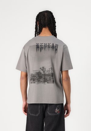 Grå bomulds t-shirt med et stort grafisk motiv på ryggen, der inkluderer tekst og ørkenbilleder, korte ærmer og en afslappet pasform.