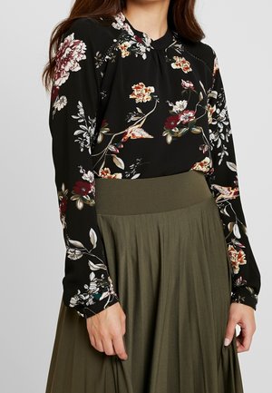 Frau trägt eine schwarze Langarmbluse mit Blumen- und Vogelmustern, kombiniert mit einem hoch taillierten olivgrünen, plissierten Rock.