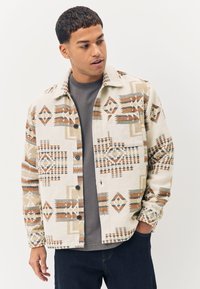 Veste crème à motifs avec des designs géométriques en marron, gris et noir. Comprend un col avec col, des fermetures à boutons et deux poches avant.