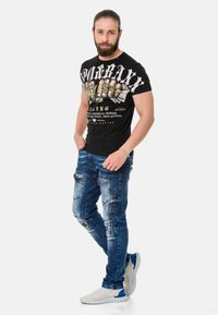 Schwarzes Grafik-T-Shirt mit fettgedrucktem Text und detaillierten Bildern, kombiniert mit abgewetzten blauen Jeans, die mehrere Taschen und leicht abgewetzte Stellen aufweisen. weiße Sneaker.