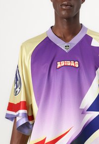 Lila és arany rövid ujjú mez, hálós anyagból, nagy "ADIDAS" logóval, jellegzetes mintákkal, valamint piros és kék színű kiemelésekkel.