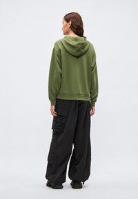 Olivovo zelená mikina s kapucňou v voľnom strihu, doplnená o voľné čierne nohavice cargo s viacerými vreckami a textúrovanou látkou.