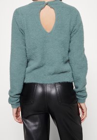 Suéter esponjoso en color verde azulado con un detalle de abertura en la espalda, combinado con pantalones de cuero negro de cintura alta que cuentan con dos bolsillos traseros.