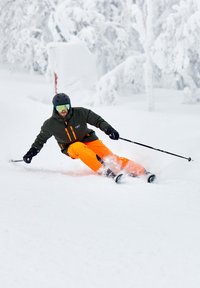 Skiër in groene jas met oranje broek, zwarte helm en grote goggles, die door diepe witte sneeuw snijdt in een besneeuwd landschap.