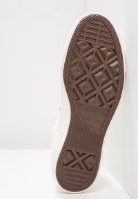 Suela de goma marrón de una zapatilla con un patrón texturizado, rodeada por una parte superior de color crema hecha de material de lona.