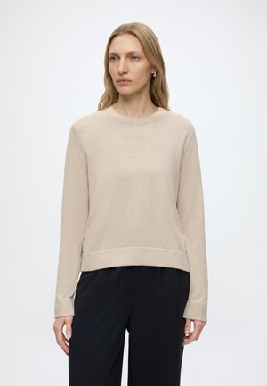 Vrouw met lang blond haar, die een beige trui met ronde hals en een zwarte broek draagt, staat tegen een effen witte achtergrond.