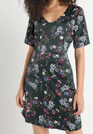 Woman wearing a dark green, short-sleeve dress with a V-neck and a multicolored floral print.