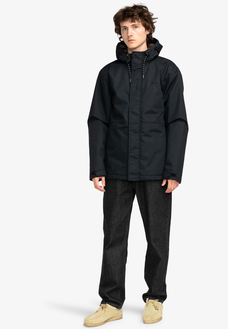 Chaqueta impermeable negra con capucha, presenta cordones ajustables y cremallera frontal. Combinada con pantalones de mezclilla oscuros y zapatos beiges.