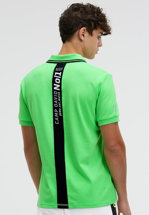 Junger Mann trägt ein hellgrünes Poloshirt mit schwarzem Streifen und weißem Text, steht mit dem Rücken zur Kamera vor einfarbigem Hintergrund.