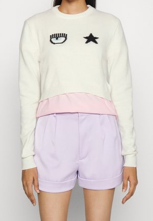Frau trägt einen kurz geschnittenen weißen Pullover mit einem Auge- und Sternenmuster, ein rosa Unterhemd und hoch taillierte fliederfarbene Shorts mit Umschlägen.