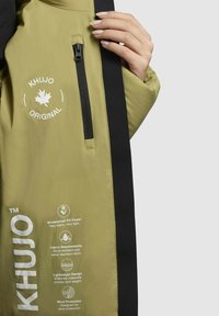 Jachetă de culoare khaki-verde cu un fermoar negru și accente negre. Are umplutură impermeabilă, material rezistent la vânt și un design ușor.