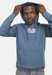 Blue hooded sweatshirt with a front pocket and drawstring hood, featuring a white logo on the chest. Cotton blend fabric, casual fit.