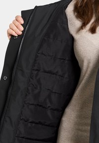 Svart vadderad jacka med en slät textur och quiltad design, i kombination med en ljusbeige ribbad topp. Dragkedja och tryckknappar synliga.