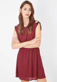 Mujer joven con cabello largo castaño, vistiendo un vestido sin mangas color burdeos con detalles de lazo negro, de pie con los brazos cruzados contra un fondo blanco.