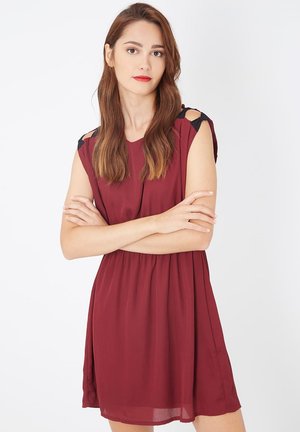 Mujer joven con cabello largo castaño, vistiendo un vestido sin mangas color burdeos con detalles de lazo negro, de pie con los brazos cruzados contra un fondo blanco.