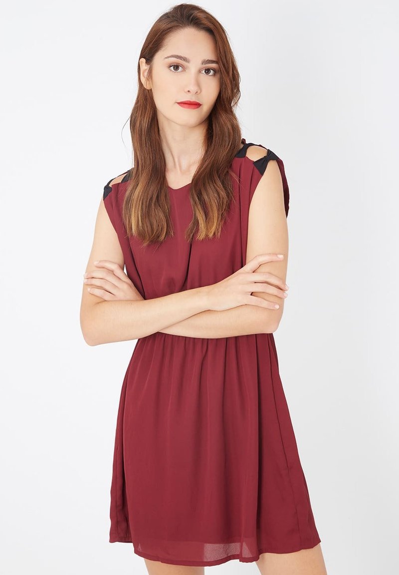 Mujer joven con cabello largo castaño, vistiendo un vestido sin mangas color burdeos con detalles de lazo negro, de pie con los brazos cruzados contra un fondo blanco.